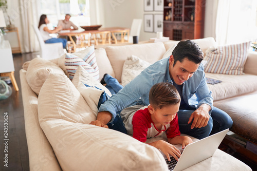 Pre-teen boy lying on sofa using laptop with his dad. Mum and grandad at the table in the background