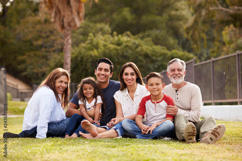 Three generation Hispanic family sitting on the grass in the park smiling to camera, selective focus Three generation Hispanic family sitting on the grass in the park smiling to camera, selective focus