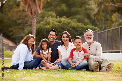 Three generation Hispanic family sitting on the grass in the park smiling to camera, selective focus