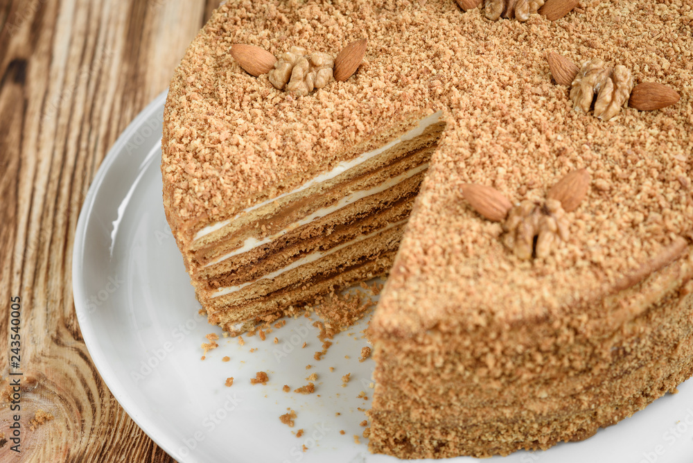 Fototapeta premium Homemade sweet layered honey cake with nuts on white plate on wooden table with bowl of honey, cinammon and badian.