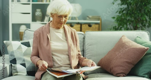 Senior Caucasian lady on retirement resting on the grey couch and flipping magazine pages in the cozy room. Inside. photo