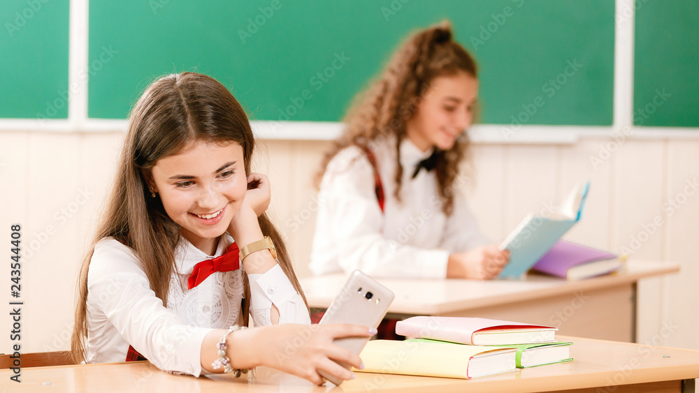 two schoolgirls are sitting at the desk in the classroom using a telephone