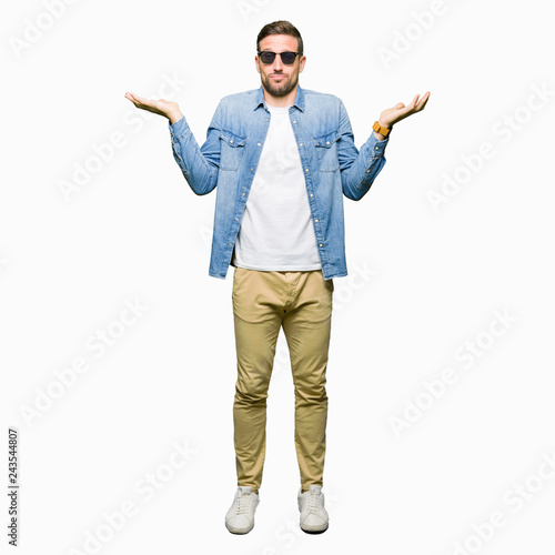Handsome man wearing fashion sunglasses clueless and confused expression with arms and hands raised. Doubt concept.