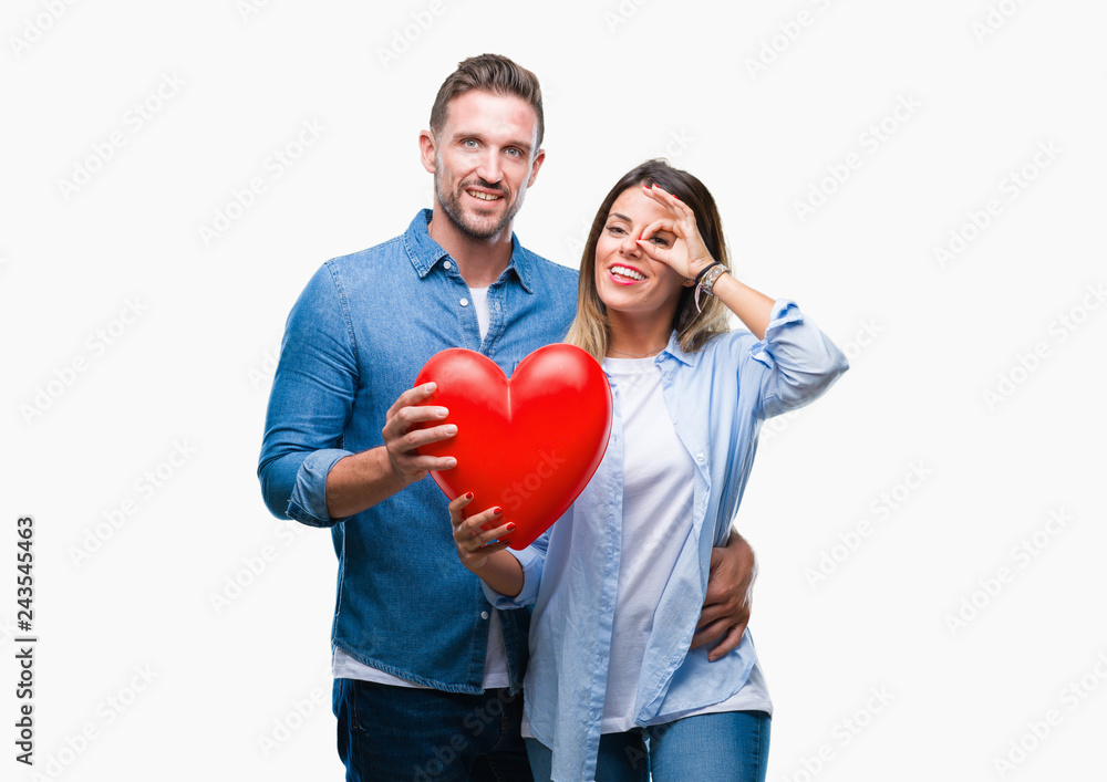 Young couple in love holding red heart over isolated background with happy face smiling doing ok sign with hand on eye looking through fingers Young couple in love holding red heart over isolated background with happy face smiling doing ok sign with hand on eye looking through fingers