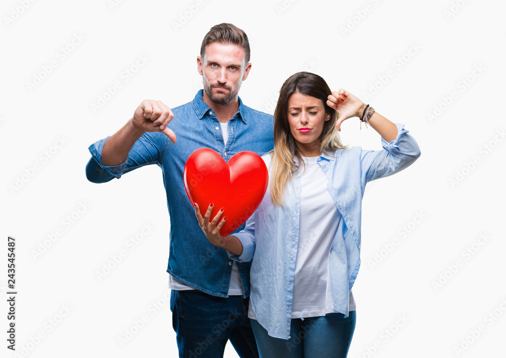 Young couple in love holding red heart over isolated background with angry face, negative sign showing dislike with thumbs down, rejection concept Young couple in love holding red heart over isolated background with angry face, negative sign showing dislike with thumbs down, rejection concept