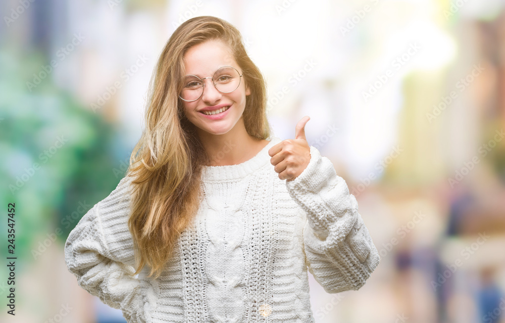 Young beautiful blonde woman wearing winter sweater and sunglasses over isolated background doing happy thumbs up gesture with hand. Approving expression looking at the camera with showing success.