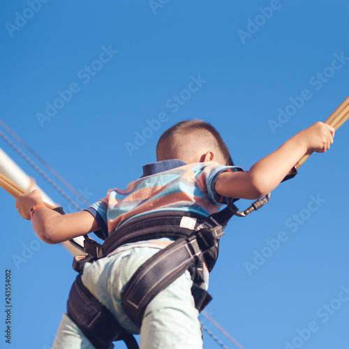 The portrait of European boy in a striped t-shirt. He is jumping on bungee trampoline high in the air against blue sky.