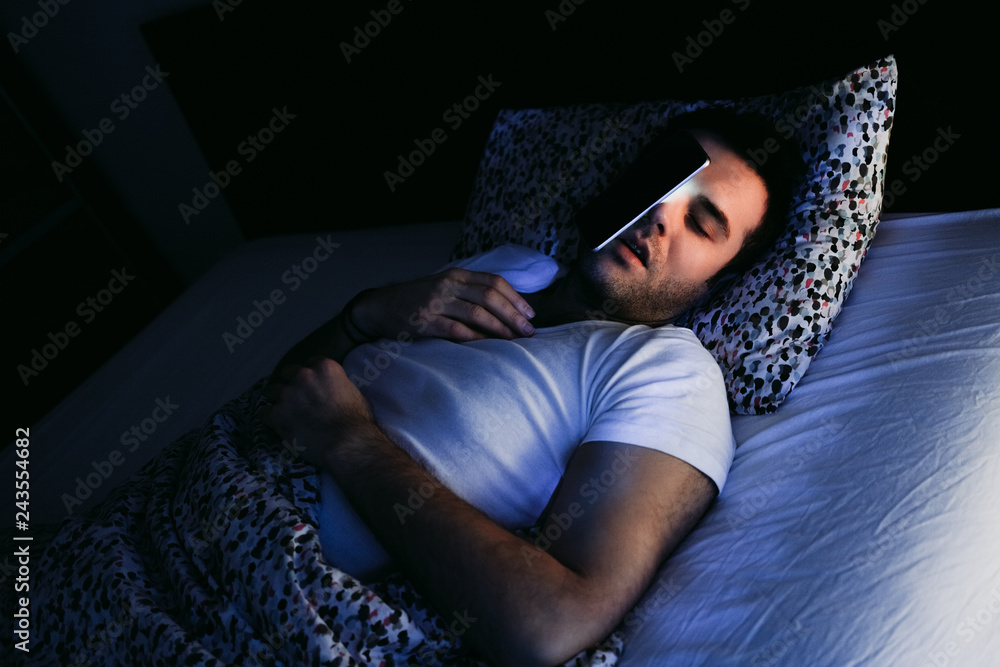 Young man using smartphone in bed at night - obrazy, fototapety, plakaty 