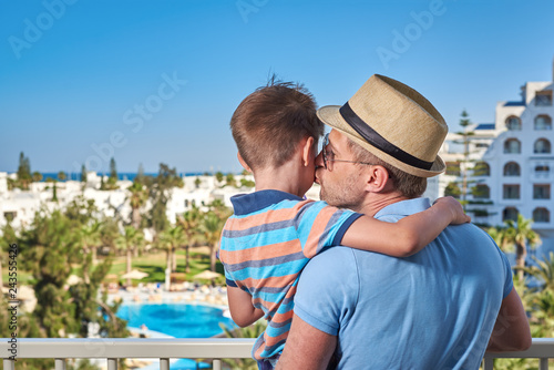 Son and dad relaxing on hotel balcony on summer holidays. Father is holding son on hands. Dad is kissing boy.
