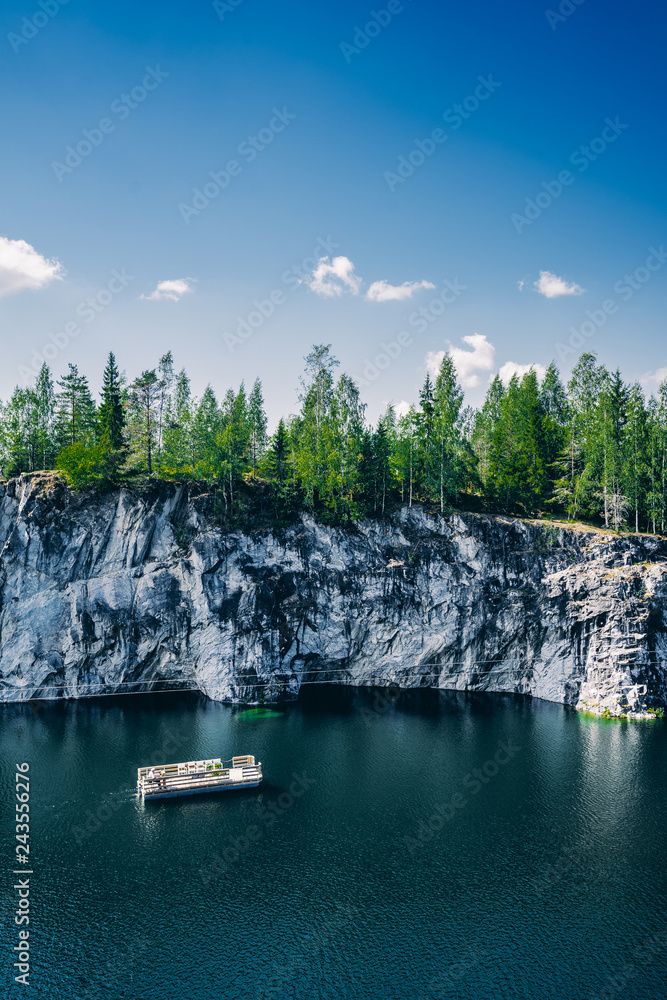 Lake with rock and forest around it, small boat floats in water. Cloudy blue sky