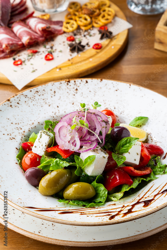 Greek salad with feta cheese, olives and herbs, served in a white plate on a table in a restaurant.