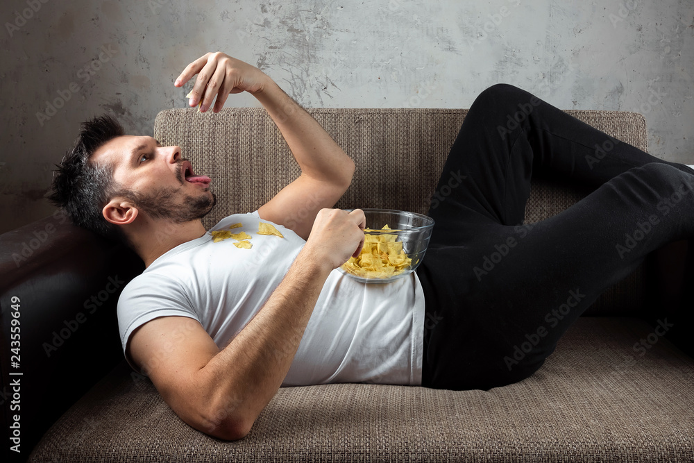 The guy in the shirt is lying on the couch, eating chips and watching a  sports channel. The concept of laziness, frustration, procrastination, the  person at home. Stock Photo | Adobe Stock