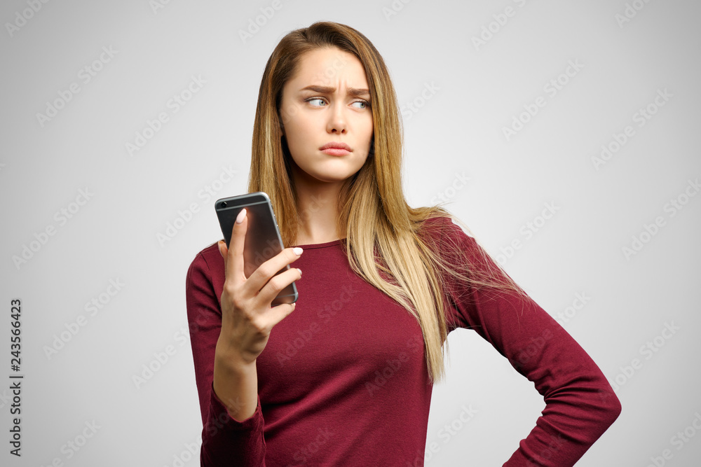 Naklejka premium Gloomy upset girl makes grimace from despair, holds smartphone, reads disapponting news, looks dejected. Isolated on white background in Studio