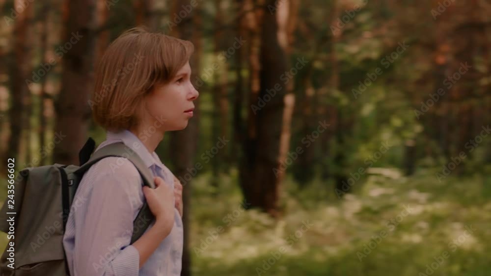 Beautiful European woman wearing backpack walking surrounded by trees in summer forest side view panning shot female traveller having good time outdoor medium shot Beautiful European woman wearing backpack walking surrounded by trees in summer forest side view panning shot female traveller having good time outdoor medium shot