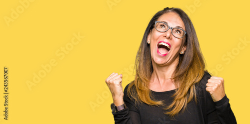 Beautiful middle age woman wearing glasses very happy and excited doing winner gesture with arms raised, smiling and screaming for success. Celebration concept.