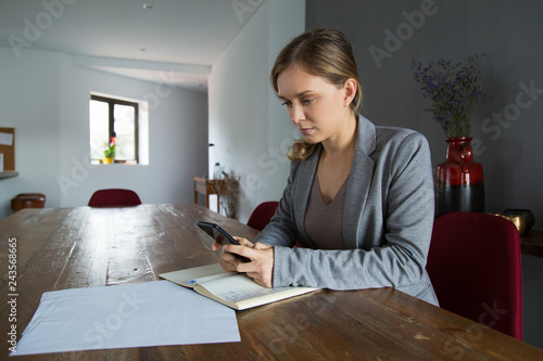 Serious professional studying documents and consulting Internet. Young woman in jacket sitting an desk with papers and notebook and using phone. Communication concept