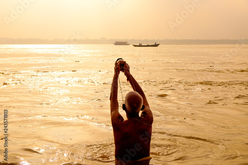evocative view of old man performing daily puja ritual on calm water of ganges river at sunrise seen from behind, with warm light photo