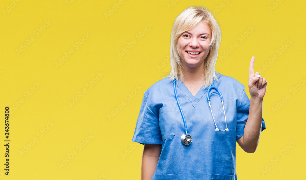Young beautiful blonde doctor woman wearing medical uniform over isolated background showing and pointing up with finger number one while smiling confident and happy. Young beautiful blonde doctor woman wearing medical uniform over isolated background showing and pointing up with finger number one while smiling confident and happy.