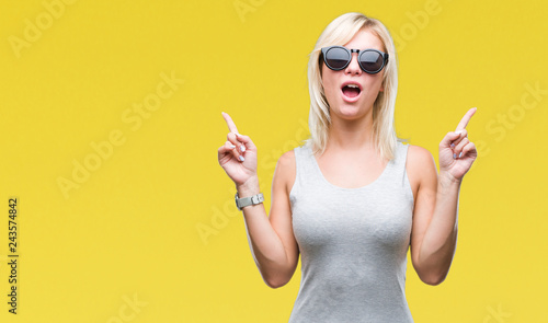 Young beautiful blonde woman wearing sunglasses over isolated background amazed and surprised looking up and pointing with fingers and raised arms.