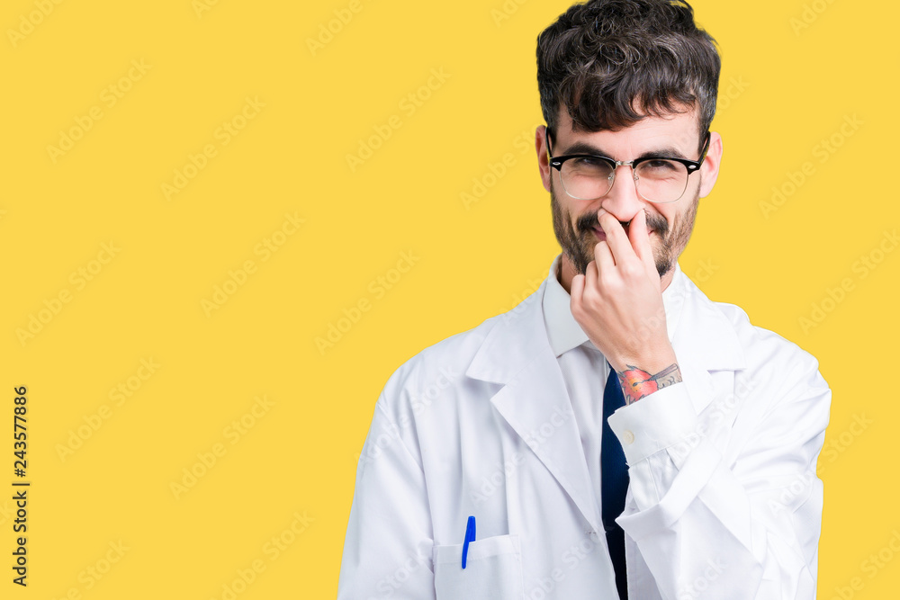 Young professional scientist man wearing white coat over isolated background smelling something stinky and disgusting, intolerable smell, holding breath with fingers on nose. Bad smells concept. Young professional scientist man wearing white coat over isolated background smelling something stinky and disgusting, intolerable smell, holding breath with fingers on nose. Bad smells concept.