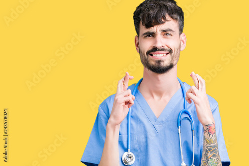 Young handsome nurse man wearing surgeon uniform over isolated background smiling crossing fingers with hope and eyes closed. Luck and superstitious concept.