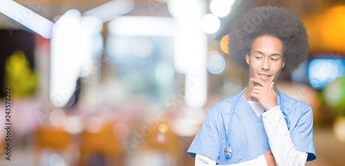Young african american doctor man with afro hair with hand on chin thinking about question, pensive expression. Smiling with thoughtful face. Doubt concept.