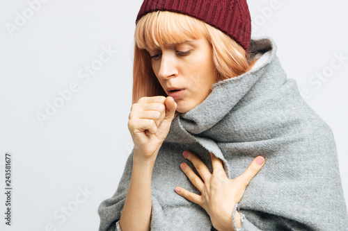 Cute sick young teen woman in red hat, wrapped in warm scarf coughing, closed eyes.Female feeling the first symptoms of illness.Bronchitis, upper respiratory infection concept.Moist cough, laryngitis photo