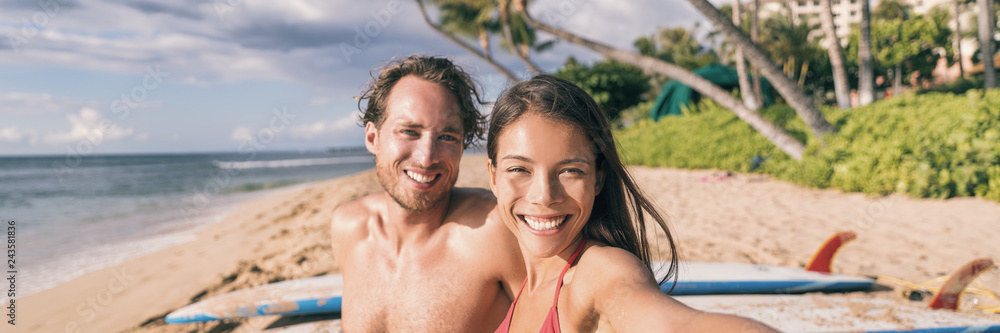 Beach vacation selfie couple. Happy young interracial friends taking photo with phone after surfing class on tropical Hawaii beach. Banner panorama. Travel vacation destination watersports. Beach vacation selfie couple. Happy young interracial friends taking photo with phone after surfing class on tropical Hawaii beach. Banner panorama. Travel vacation destination watersports.