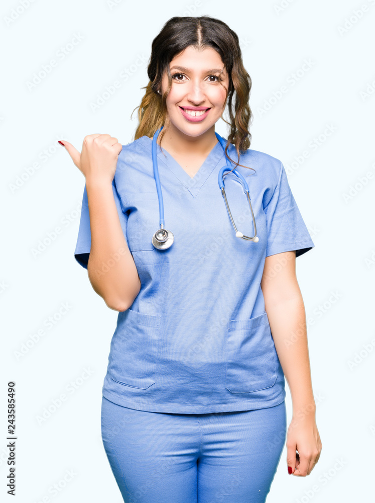 Young adult doctor woman wearing medical uniform smiling with happy face looking and pointing to the side with thumb up. Young adult doctor woman wearing medical uniform smiling with happy face looking and pointing to the side with thumb up.