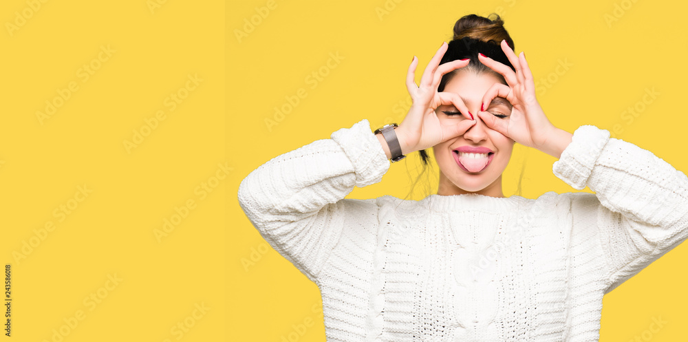 Young beautiful woman wearing winter sweater doing ok gesture like binoculars sticking tongue out, eyes looking through fingers. Crazy expression. Young beautiful woman wearing winter sweater doing ok gesture like binoculars sticking tongue out, eyes looking through fingers. Crazy expression.