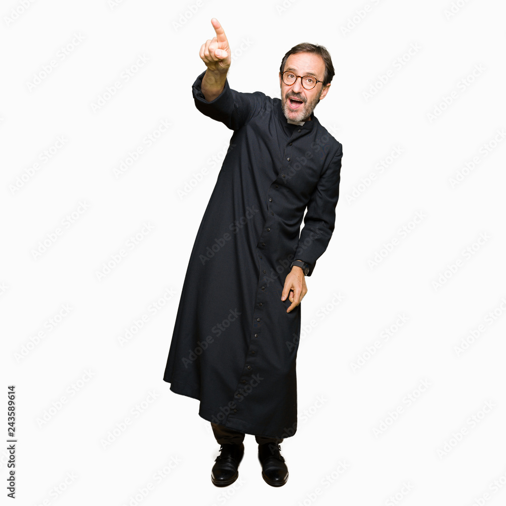 Middle age priest man wearing catholic robe Pointing with finger surprised ahead, open mouth amazed expression, something in front