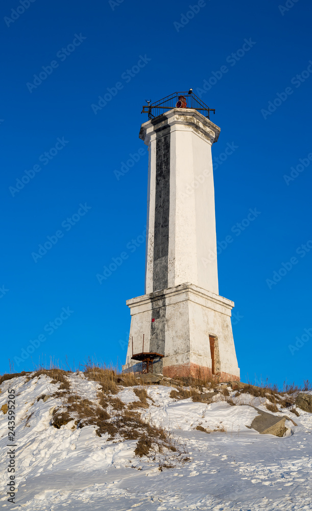 Leading beacon for marine navigation. High white lighthouse on the snow-covered shore against a blue sky. Cold winter. Vertical orientation. Magadan, Nagaev Bay, Sea of Okhotsk, Far East of Russia.