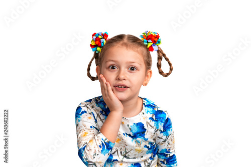 Studio waist up portrait of a little girl wearing shirt with a print and with two pigtails and color bows on a gray background. She is holding her cheek with her palm and is surprised at something