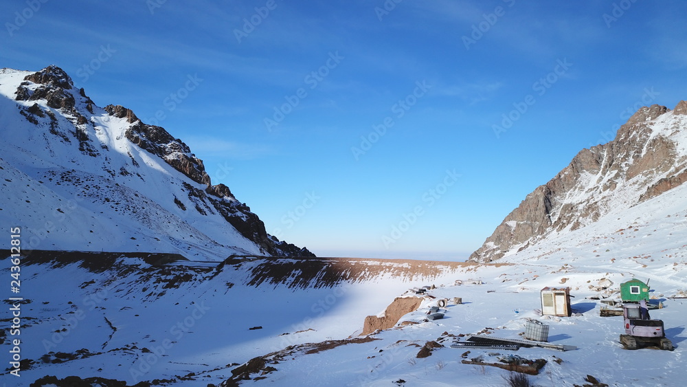 Construction equipment in the snowy mountains. Small house, tractor, equipment for snow the dam. Among mountains and rocks. View of the blue sky and white clouds. Winter.