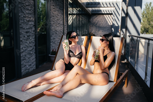 Two girls or women in vacation, Asian and Caucasian, in tropical garden at hotel pool tanning being served drinks or cocktails