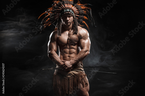 American Indian Apache warrior chief  in traditional clothing and feathered headdress with weapon. Indian chieftain of the tribe with muscled strength body on smoke dark background. photo
