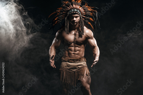 American Indian Apache warrior chief in traditional clothing and feathered headdress with weapon. Indian chieftain of the tribe with muscled strength body on smoke dark background.