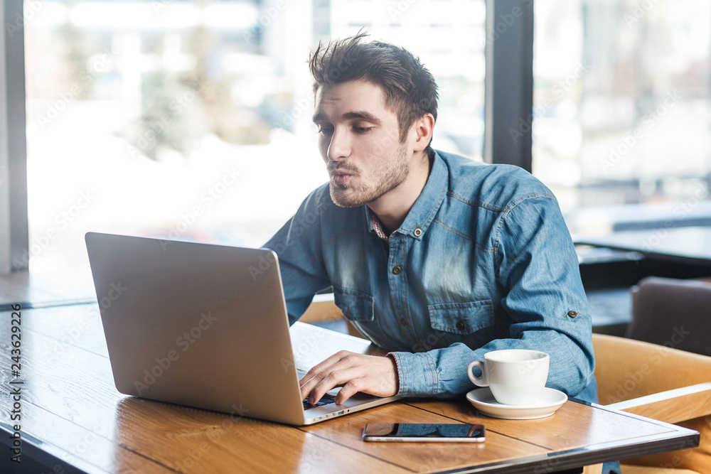 Side view portrait of lovely bearded young handsome man in blue jeans shirt are sitting alone in cafe and making video call sending air kiss to his girlfriend on laptop. Side view portrait of lovely bearded young handsome man in blue jeans shirt are sitting alone in cafe and making video call sending air kiss to his girlfriend on laptop.
