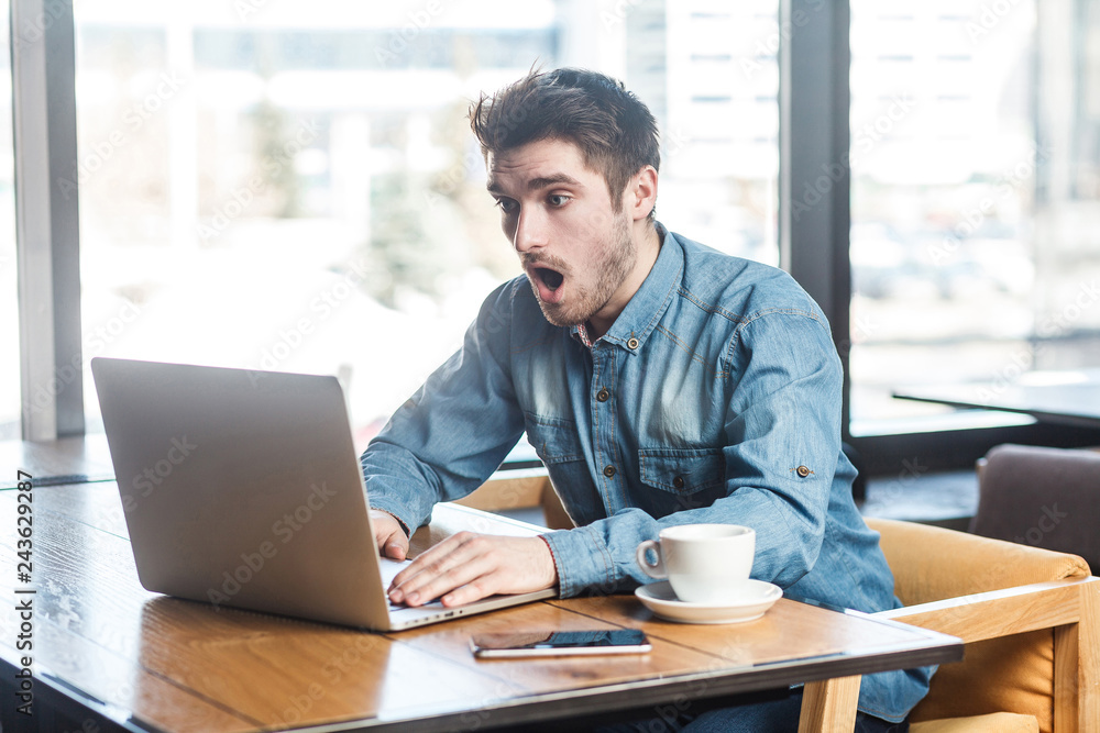 Don`t believe! Side view portrait of emotional shocked young businessman in blue jeans shirt are sitting in cafe, reading news and remotely working with suprised big eyes looking to the laptop. Don`t believe! Side view portrait of emotional shocked young businessman in blue jeans shirt are sitting in cafe, reading news and remotely working with suprised big eyes looking to the laptop.