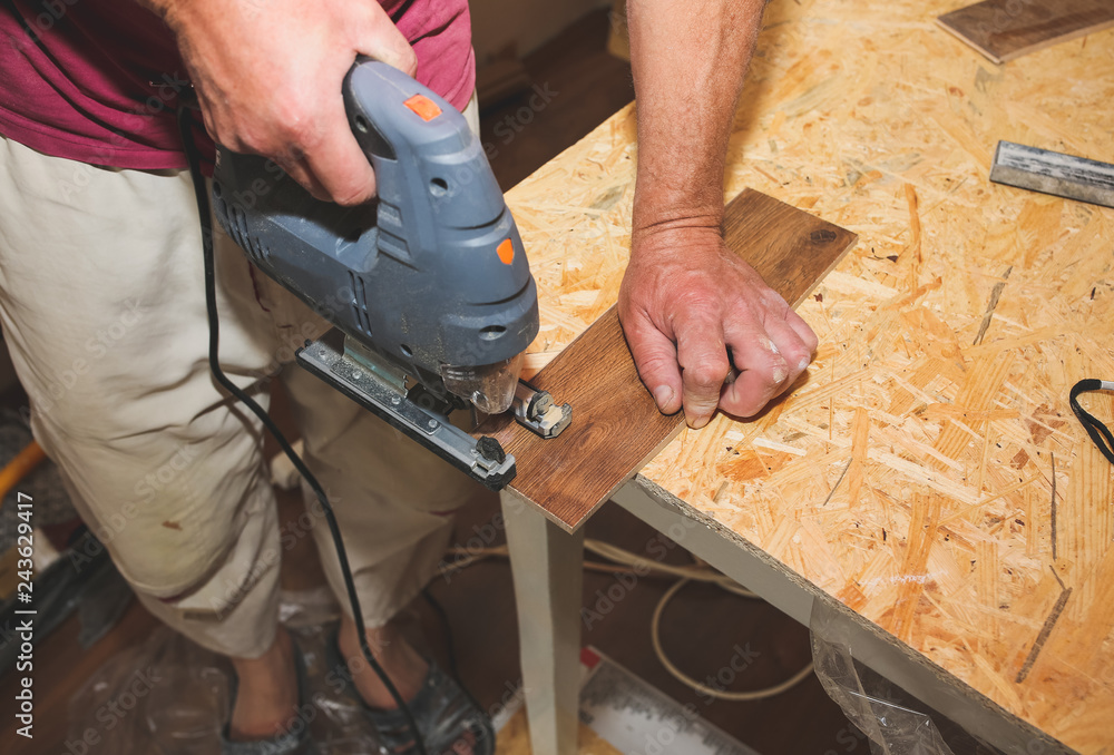 Worker is holding in hands power jigsaw and cutting laminate flooring in apartment. Maintenance repair works renovation in the flat with scroll saw. Restoration of wooden parquet floor planks indoors.