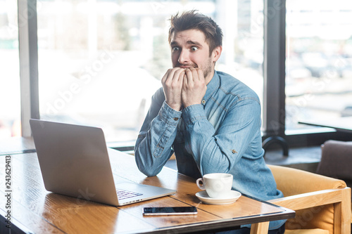 Wall Mural Scared! Side view portrait of emotional nervous young businessman in blue jeans shirt are sitting in cafe, working onlone and nail biting himself cause made big mistake in the laptop