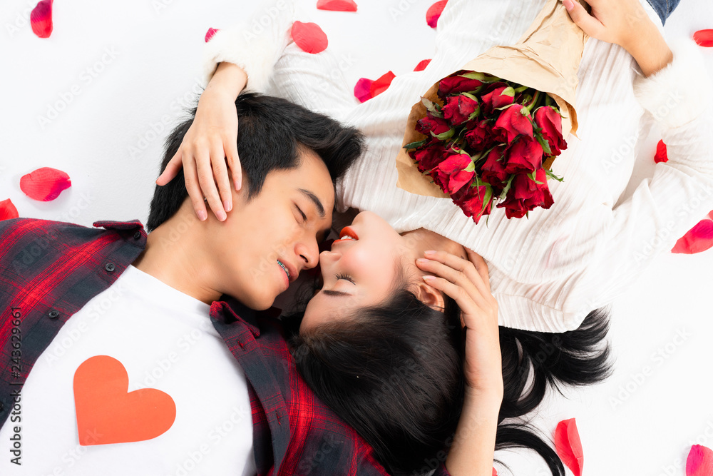 Top view of happy young couple looking at each other and smiling while lying on white background floor. They is holding a red paper heart, and Rose petals fall full on the White Background Top view of happy young couple looking at each other and smiling while lying on white background floor. They is holding a red paper heart, and Rose petals fall full on the White Background