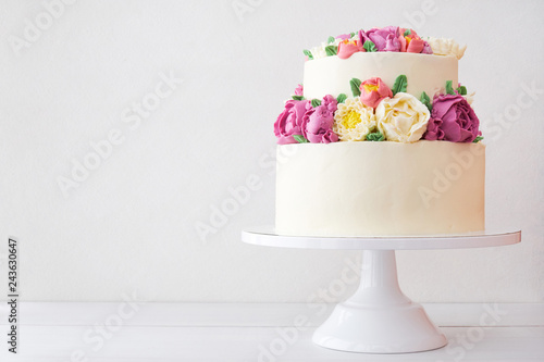 Two-tiered white wedding cake decorated with color cream flowers on a white wooden background.