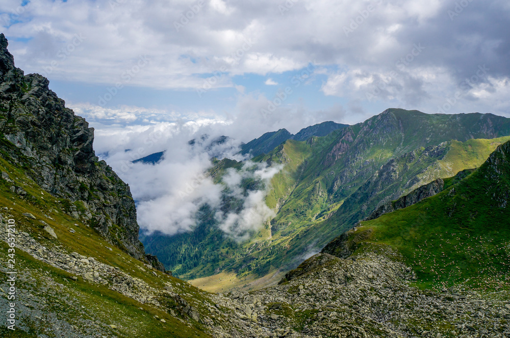 Dramatic mountain landscape with low clouds and sheer rocks covered in green grass in Fagaras Mountains, Carpathian range, Romania on a summer day.