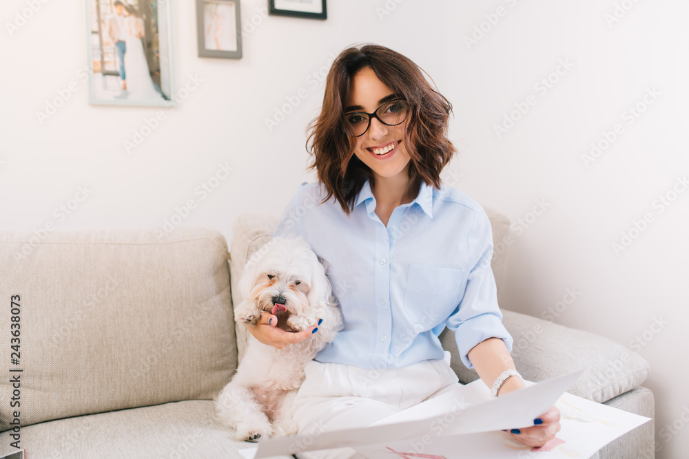 A brunette young girl in a blue shirt is sitting on the sofa in studio. She hugs white small dog and holds some sketches on her knees. She is smiling to the camera. A brunette young girl in a blue shirt is sitting on the sofa in studio. She hugs white small dog and holds some sketches on her knees. She is smiling to the camera.