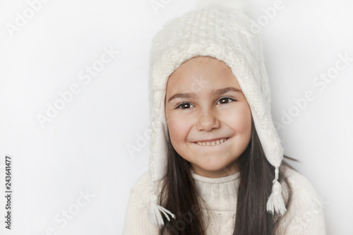 Happy cute child girl in a white winter hat and a white knitted sweater laughs on a white background. Real positive emotions. Smiling beautiful face. 