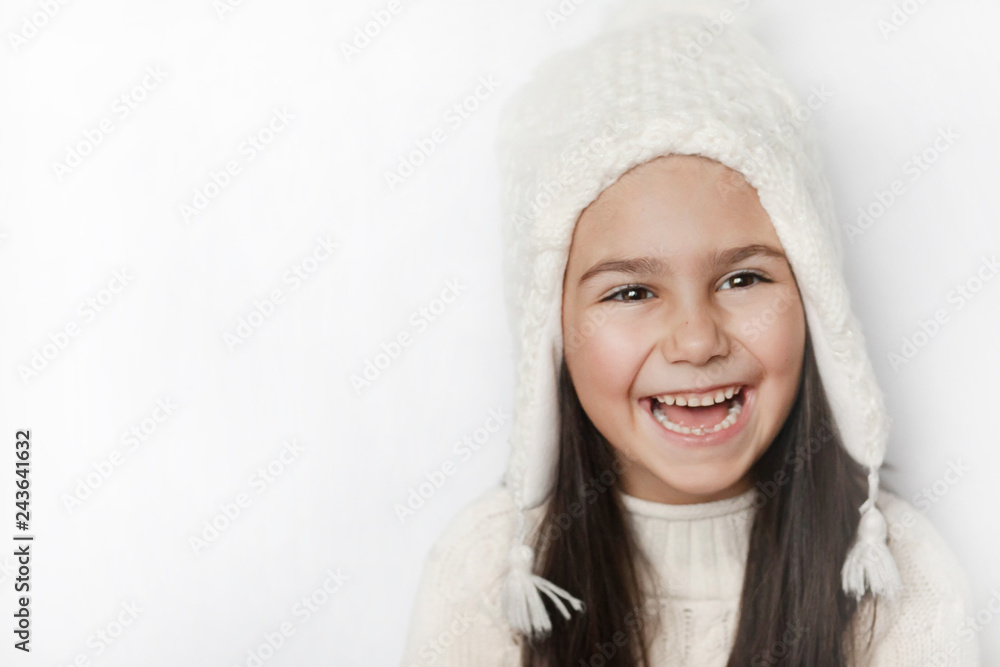 Obraz premium Happy cute child girl in a white winter hat and a white knitted sweater laughs on a white background. Real positive emotions. Smiling beautiful face. 