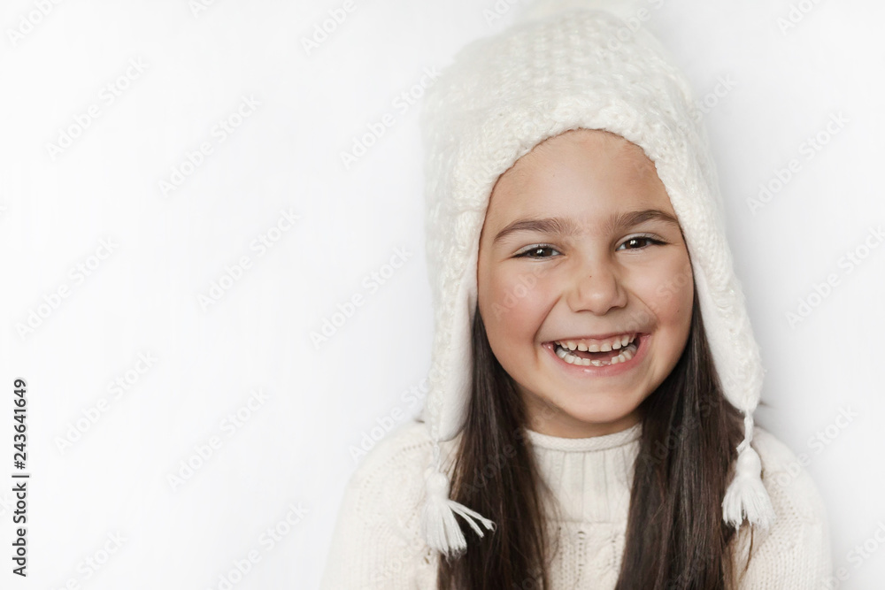 Obraz premium Happy cute child girl in a white winter hat and a white knitted sweater laughs on a white background. Real positive emotions. Smiling beautiful face. 