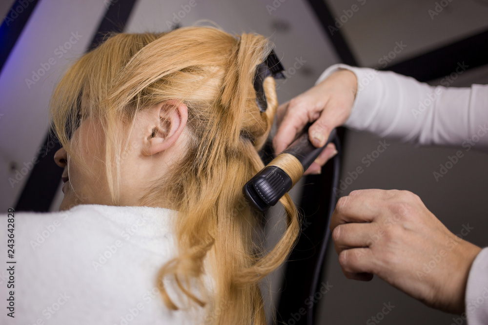 Fototapeta premium Close-up photo professional hairdresser uses flat iron for making beautiful curls, blonde hair. Client sitting in the chair