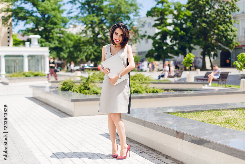 Pretty brunette girl in gray dress and vinous heels is standing in park in the city. She holds laptop and looks shy.